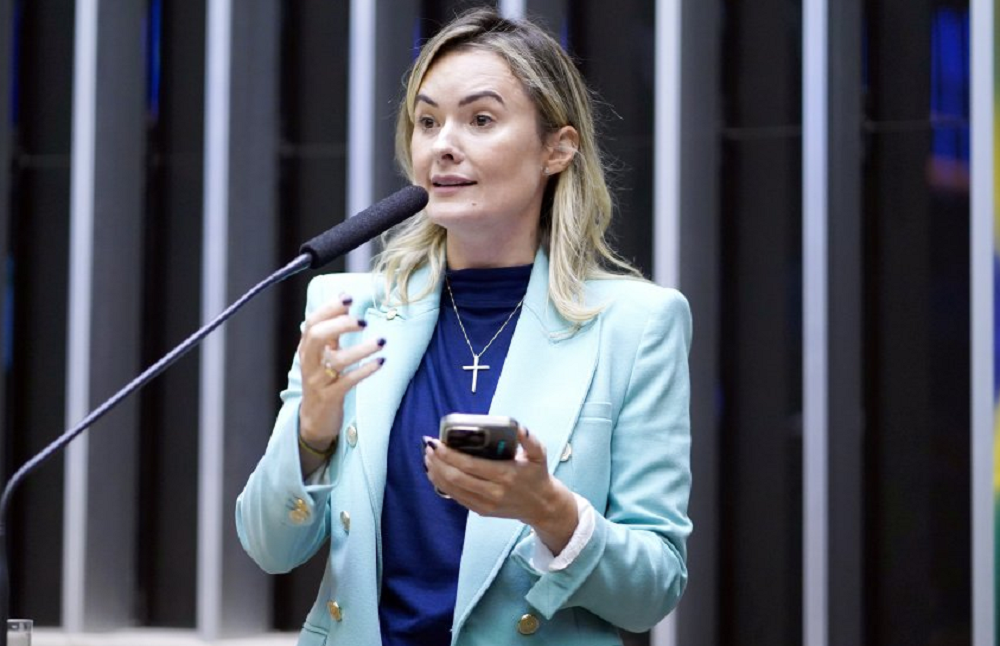 A deputada federal Júlia Zanatta (PL-SC) de roupa social azul, falando em microfone e segurando celular, de cabelo solto