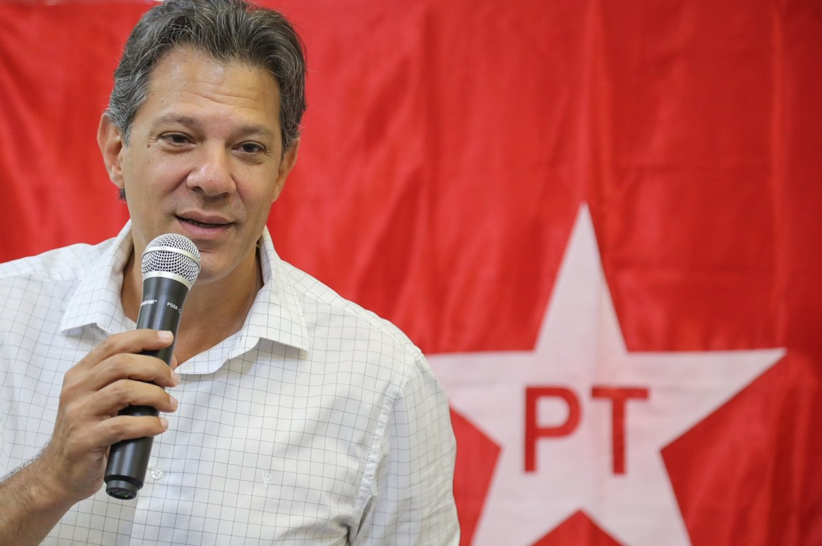O ministro da Fazenda, Fernando Haddad, de roupa branca, falando em microfone com bandeira do PT ao fundo