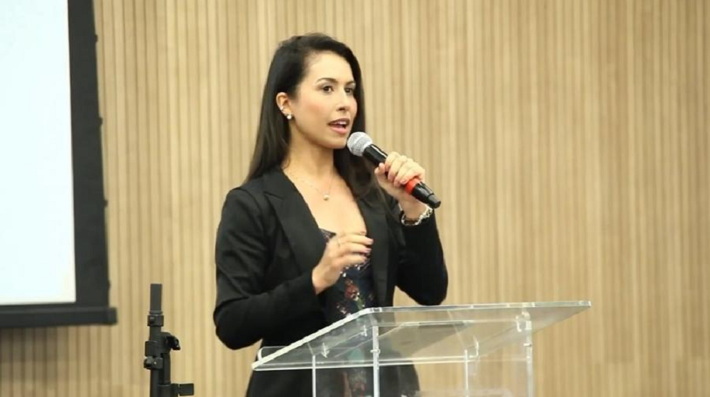 Ludmila Lins Grilo de roupa preta, em pé, falando em microfone, de cabelo solto