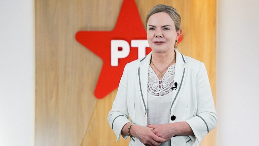 Gleisi Hoffmann de roupa branca e cabelo preso, perto de parede com símbolo do PT