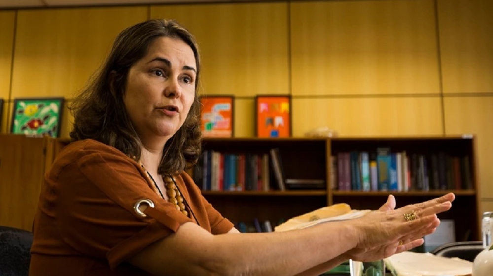 A procuradora da República Eugênia Augusta Gonzaga, de roupa caramelo, perto de livros, falando e gesticulando, séria