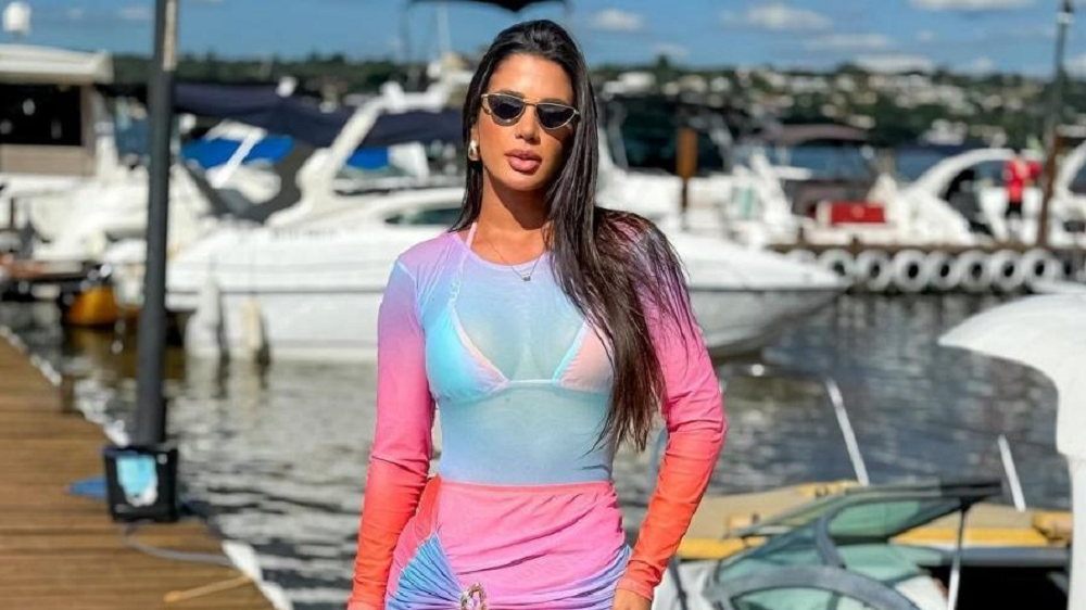 A influenciadora digital Aline Ferreira posando em píer, com roupa colorida e óculos escuros, séria, de cabelo solto