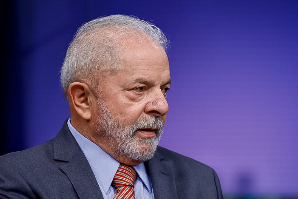 Lula de perfil, sério, de roupa social, em fundo roxo