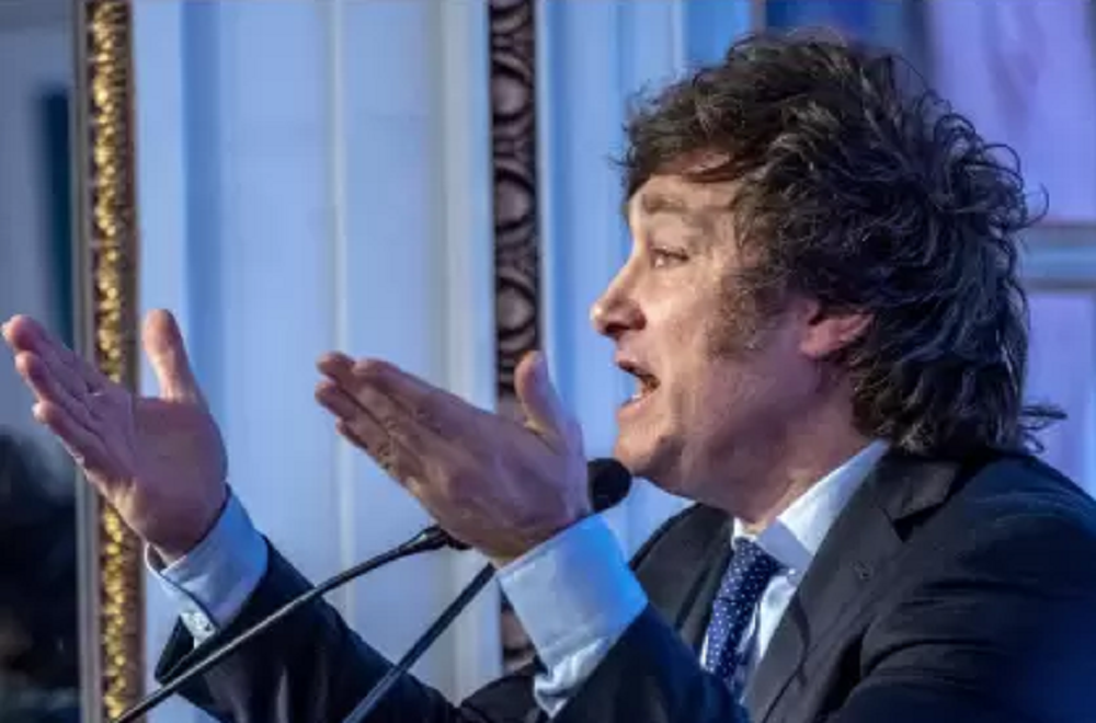 Javier Milei de perfil, falando em microfone e gesticulando, com raiva