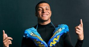 O capitão da seleção francesa de futebol, Mbappé, de camisa preta de frio, com duas chuteiras azuis penduradas nos ombros, sorrindo e apontando pra cima