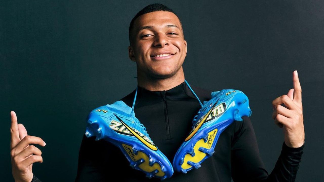 O capitão da seleção francesa de futebol, Mbappé, de camisa preta de frio, com duas chuteiras azuis penduradas nos ombros, sorrindo e apontando pra cima