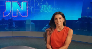 Mônica Teixeira na bancada do Jornal Nacional, de roupa coral e cabelo solto, com sorriso tímido