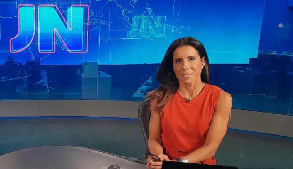 Mônica Teixeira na bancada do Jornal Nacional, de roupa coral e cabelo solto, com sorriso tímido