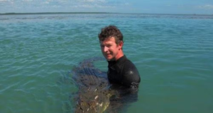 Adam Britton sorrindo para foto na água com crocodilo