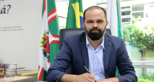 O secretário de Educação do Paraná, Roni Miranda, sério, escrevendo em papel, com bandeiras ao fundo