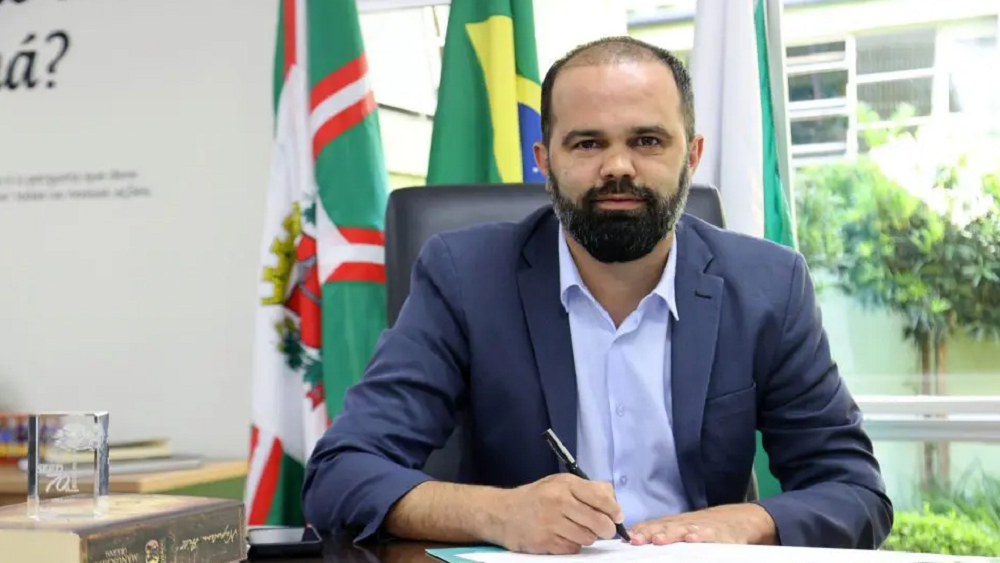 O secretário de Educação do Paraná, Roni Miranda, sério, escrevendo em papel, com bandeiras ao fundo