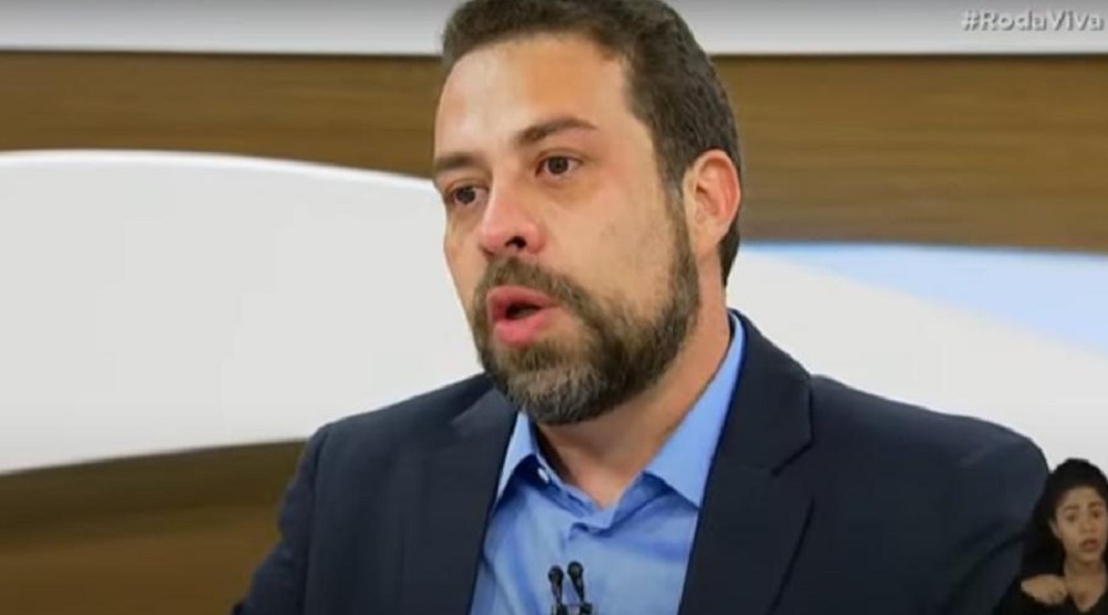 Guilherme Boulos de roupa social azul, sem olhar para a câmera, falando, no Roda Viva