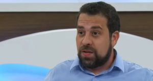 Guilherme Boulos no cenário do Roda Viva, de camisa social azul, falando, sem olhar para a câmera