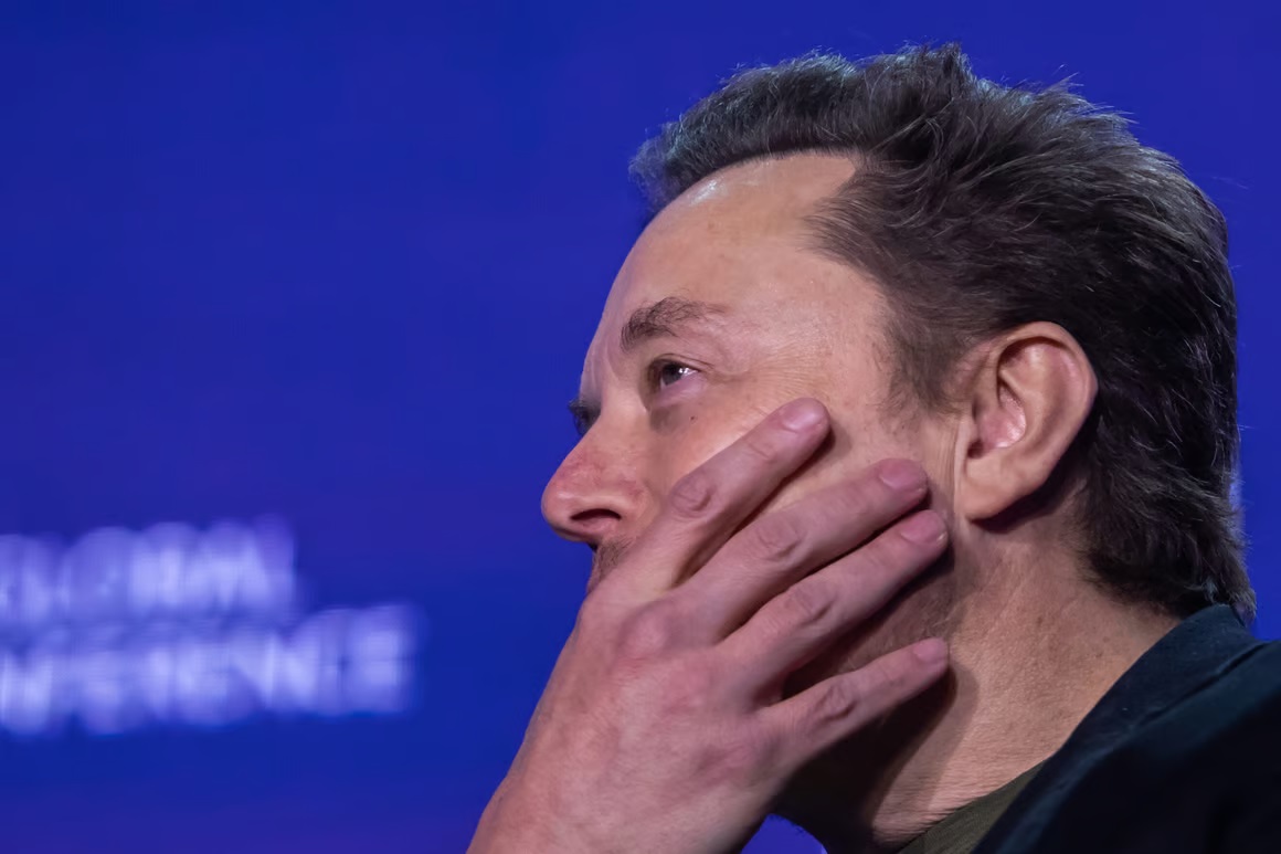 Elon Musk de perfil, com mão cobrindo parte do rosto, sério