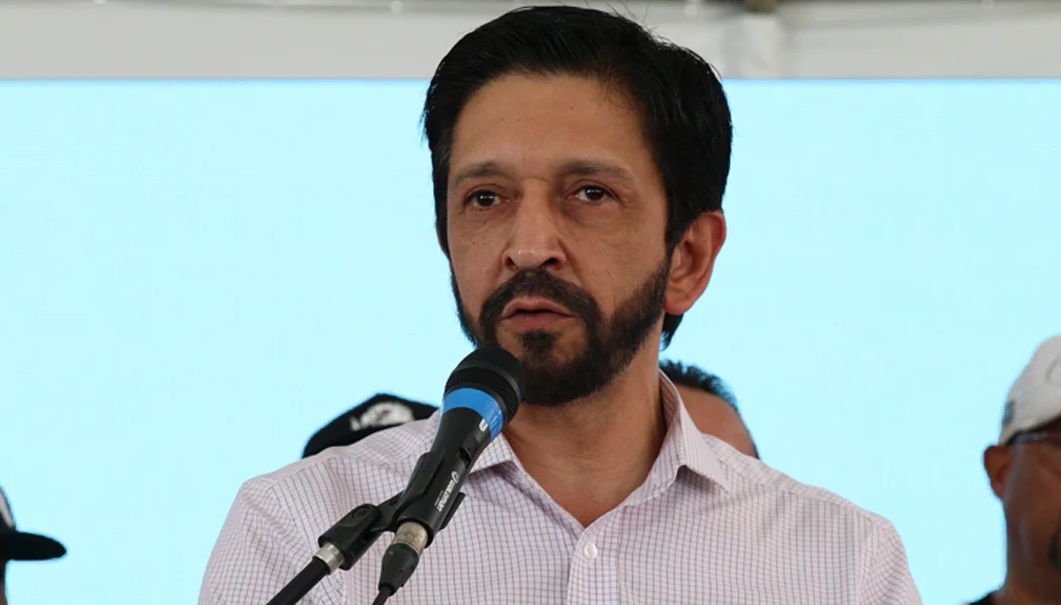 O prefeito Ricardo Nunes (MDB) com expressão assustada e camisa branca, falando em microfone