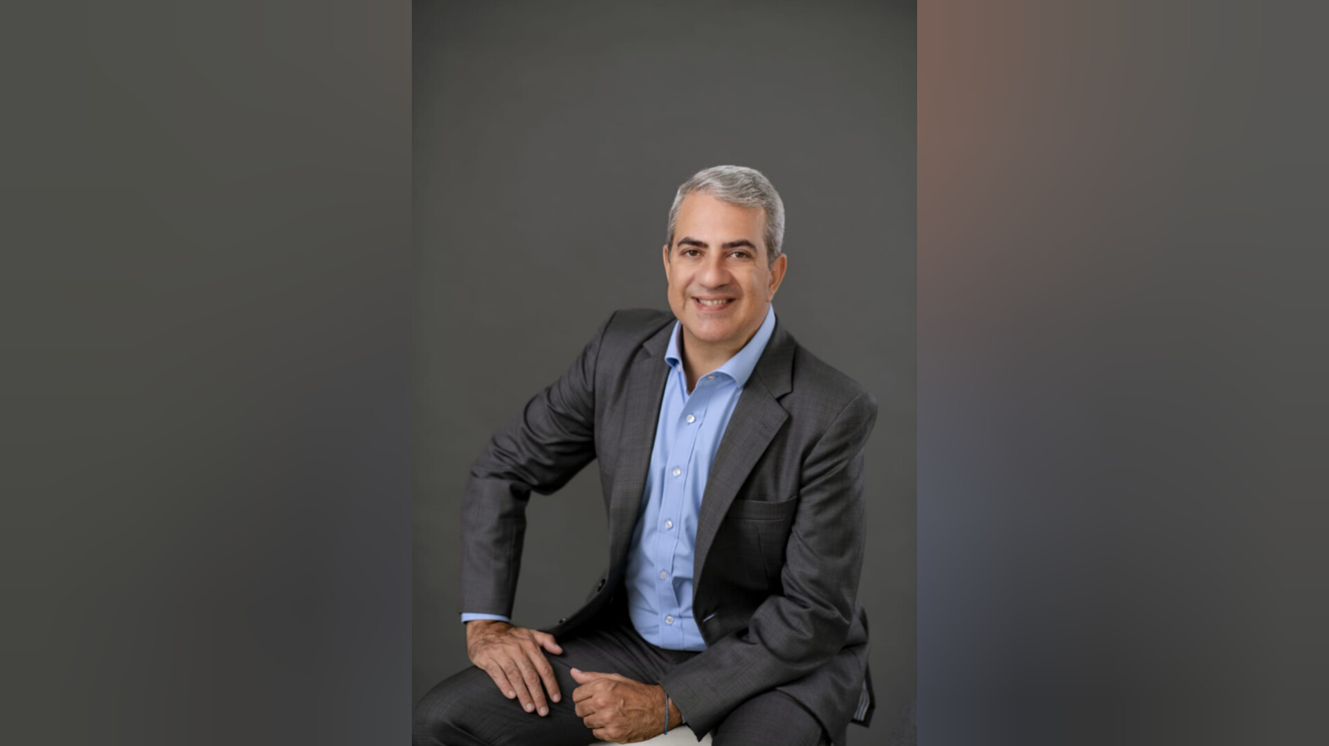 Wilson Pedroso Júnior, também conhecido como Wilsinho, de roupa social cinza, sorrindo, olhando para a câmera, sentado