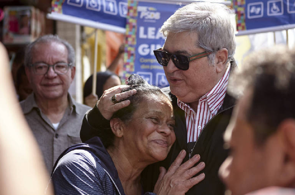 Datena (PSDB) abraçando fã na Vila Nova Cachoeirinha, em São Paulo, ele de óculos escuros e falando, ela sorrindo, rodeados por pessoas
