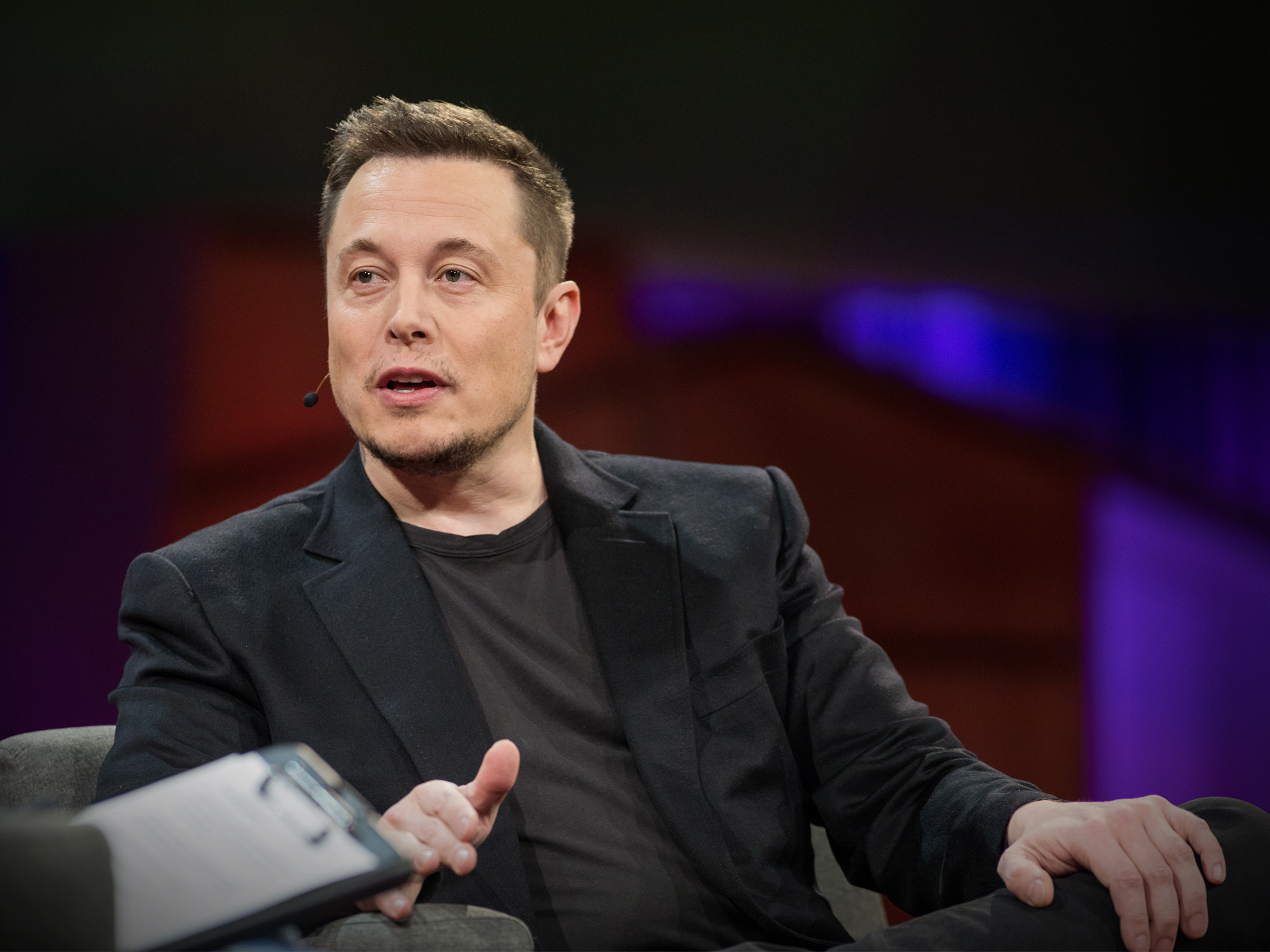 Elon Musk de roupa social preta, falando sem olhar para a câmera