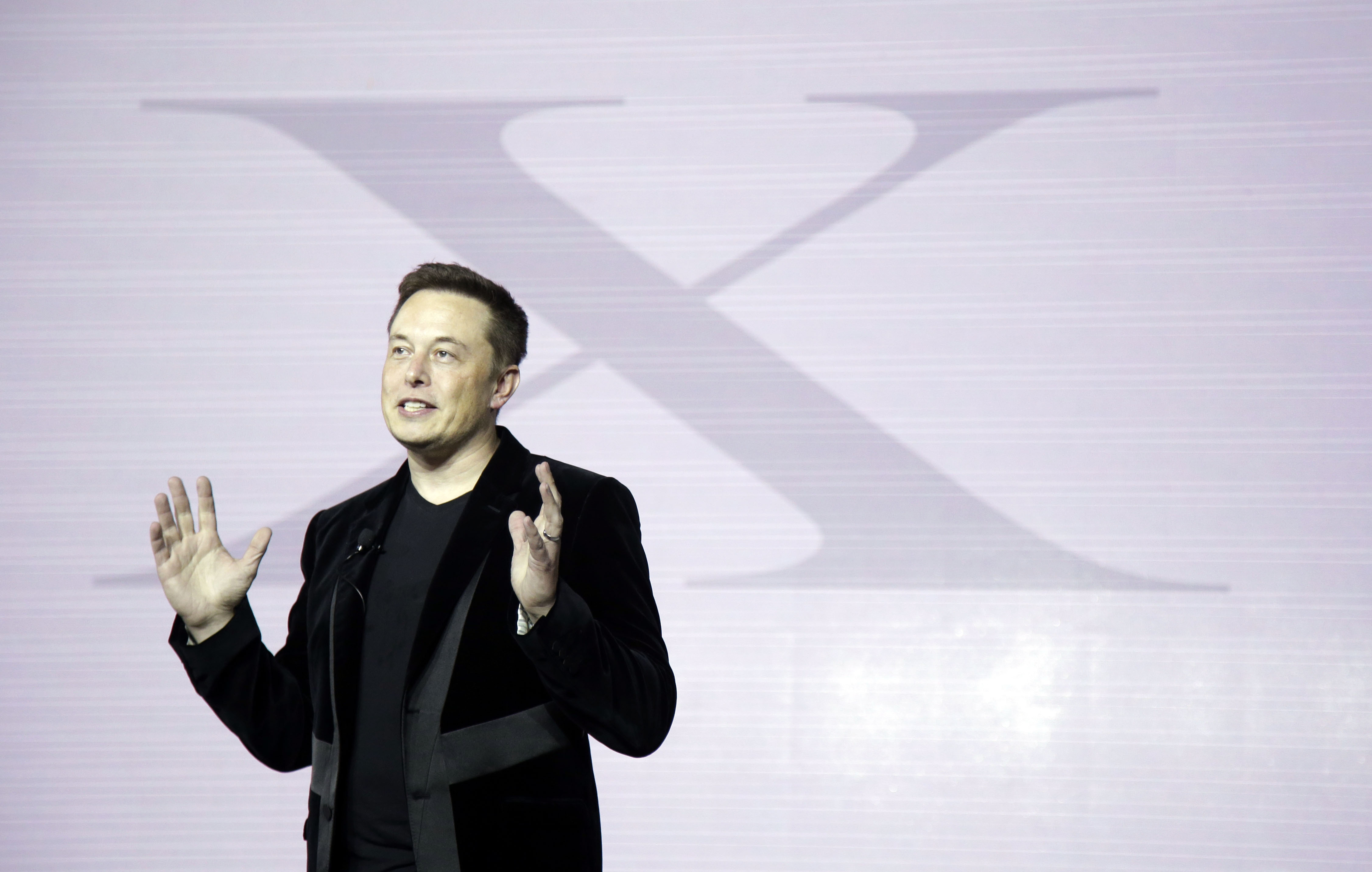 Elon Musk olhando para o lado, falando e gesticulando, com a letra X em cinza ao fundo