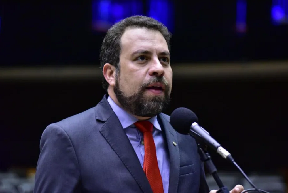 Guilherme Boulos falando em microfone, sério, sem olhar para a câmera