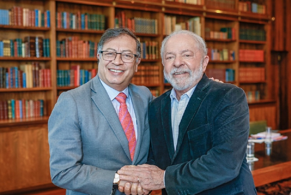 Gustavo Petro e Lula em aperto de mãos, sorrindo e posando para foto com estantes de livros ao fundo