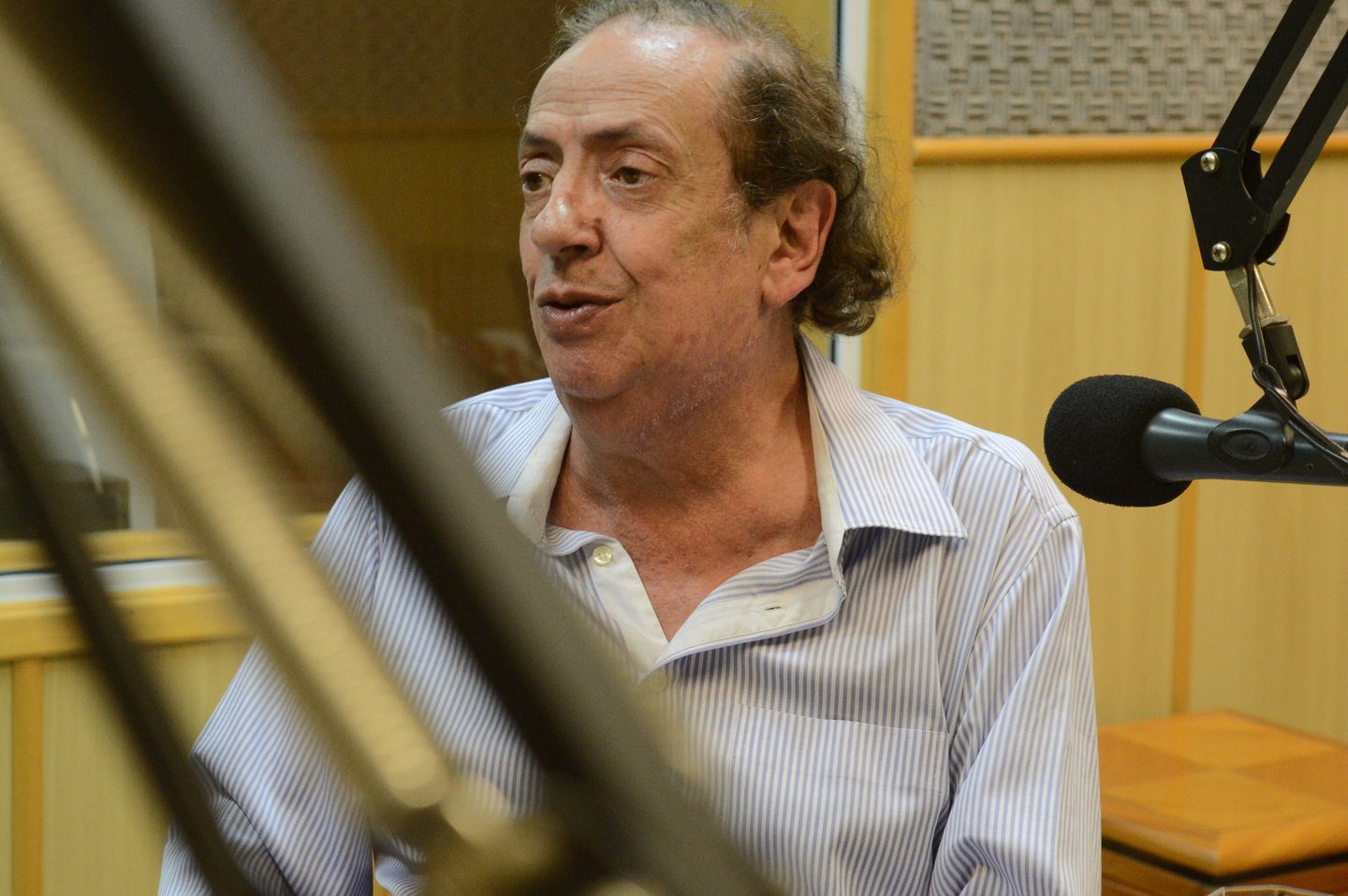 Marcos Oliveira de perfil, de camisa social clara, falando em microfone em estúdio