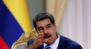 Nicolás Maduro falando e apontando pra frente, com bandeira da Venezuela ao fundo, sério, sem olhar para a câmera