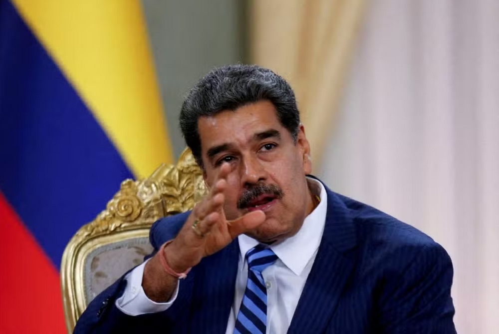 Nicolás Maduro falando e apontando pra frente, com bandeira da Venezuela ao fundo, sério, sem olhar para a câmera
