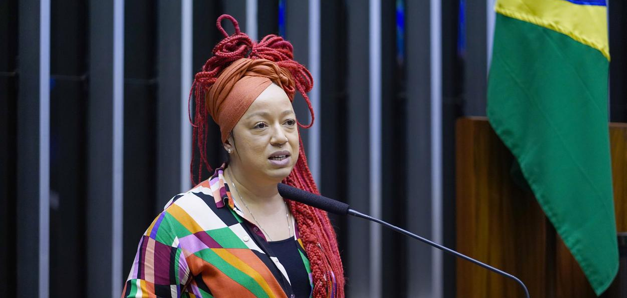 A deputada federal Daiana Santos (PCdoB-RS) com expressão de tristeza, falando em microfone, sem olhar para a câmera