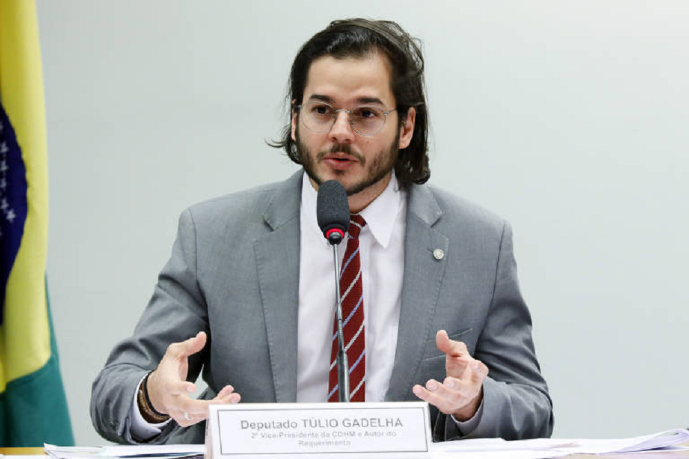 O deputado federal Túlio Gadêlha (Rede-PE) falando e gesticulando, de roupa social cinza e óculos, sem olhar para a câmera