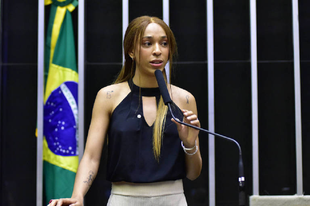 A deputada Érika Hilton (PSOL) de roupa preta e cabelo solto, falando em microfone, séria