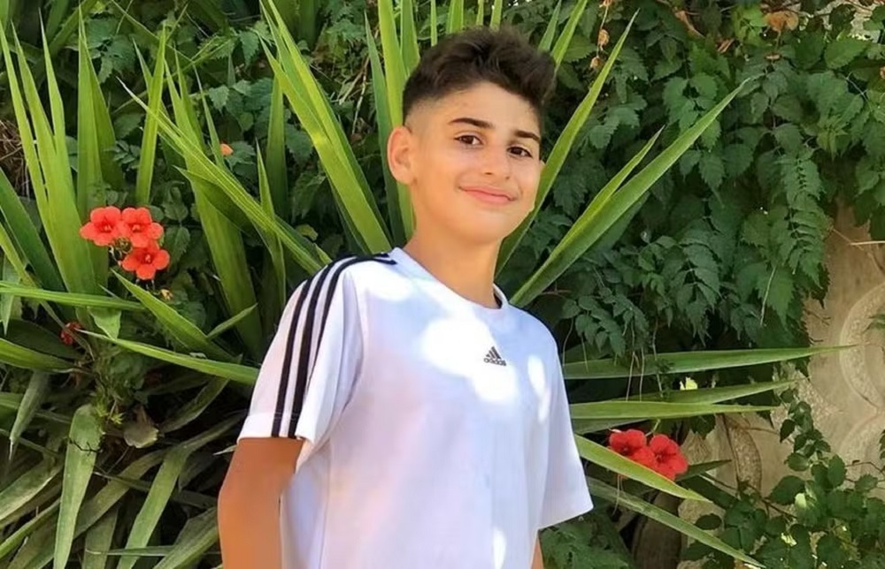 Ali Kamal Abdallah de camiseta branca com detalhes pretos e sorriso tímido para a câmera, posando perto de plantas