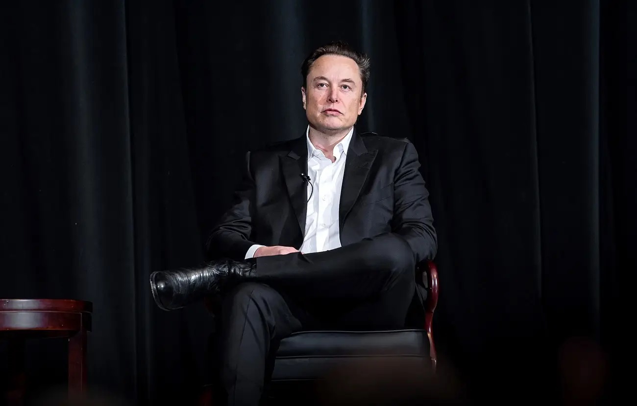 Elon Musk sério, de roupa social, sentado