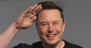 Elon Musk sorrindo, de camiseta preta, fazendo sinal de continência