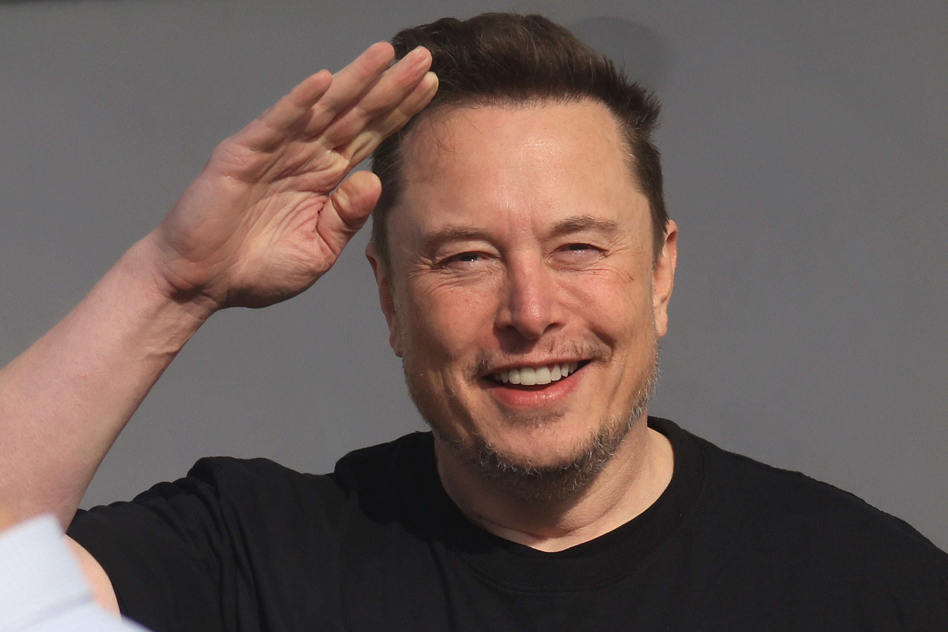Elon Musk sorrindo, de camiseta preta, fazendo sinal de continência