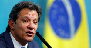 O Ministro da Fazenda Fernando Haddad com expressão séria, falando, com bandeira do Brasil ao fundo