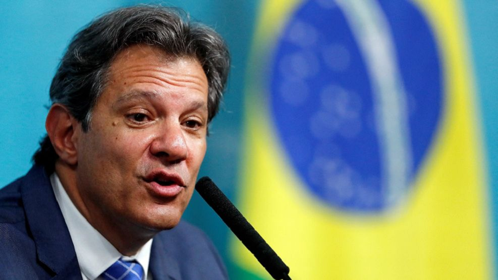 O Ministro da Fazenda Fernando Haddad com expressão séria, falando, com bandeira do Brasil ao fundo