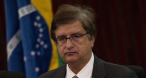 O procurador-geral da República, Paulo Gonet, sério, de óculos, com bandeira do Brasil ao fundo