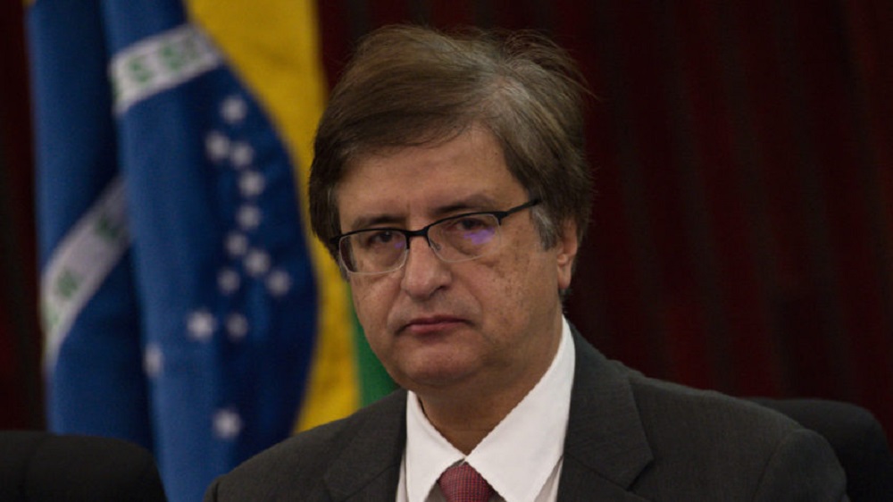 O procurador-geral da República, Paulo Gonet, sério, de óculos, com bandeira do Brasil ao fundo