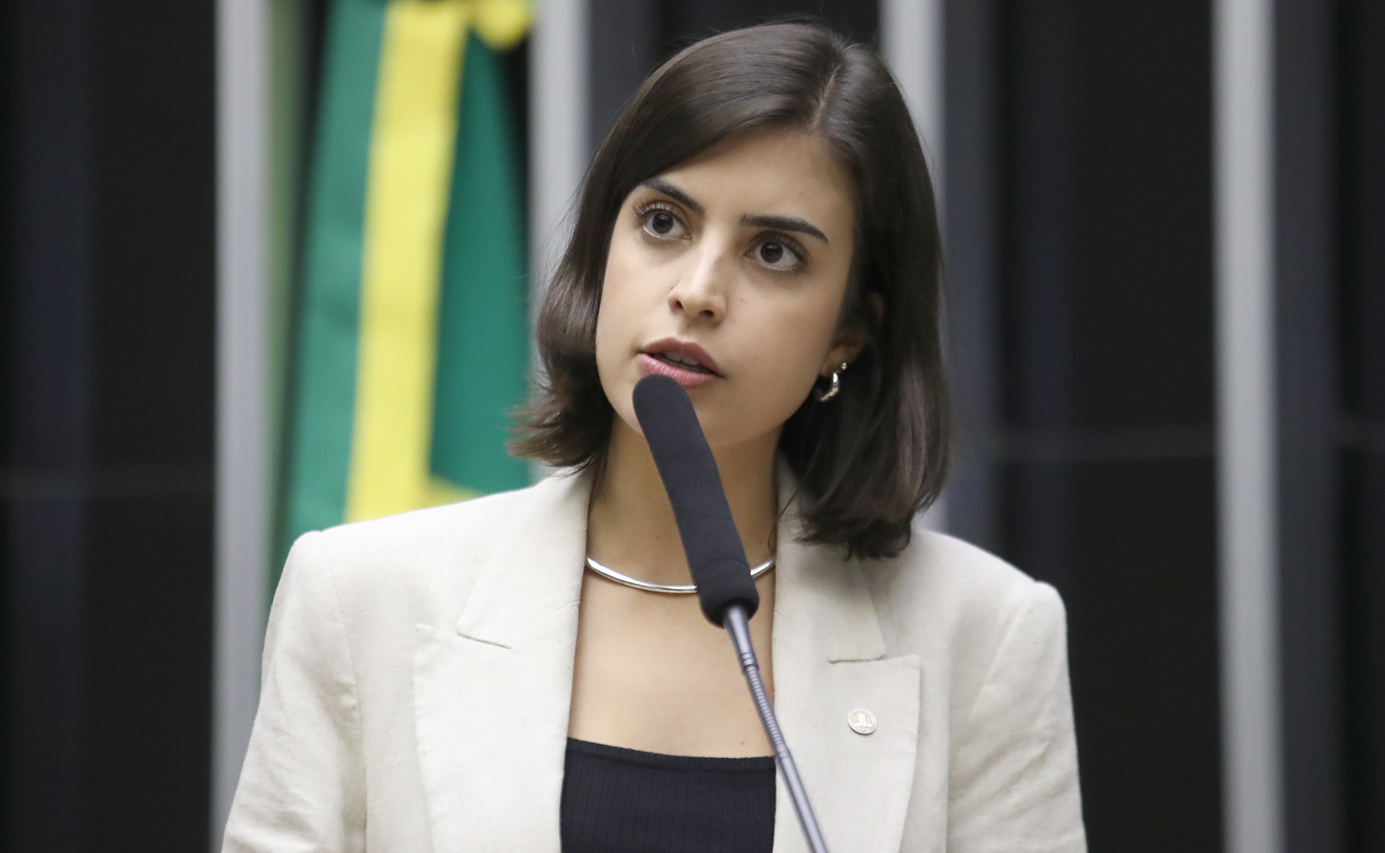 Tabata Amaral falando em microfone na Câmara dos Deputados, séria, de roupa bege e preta
