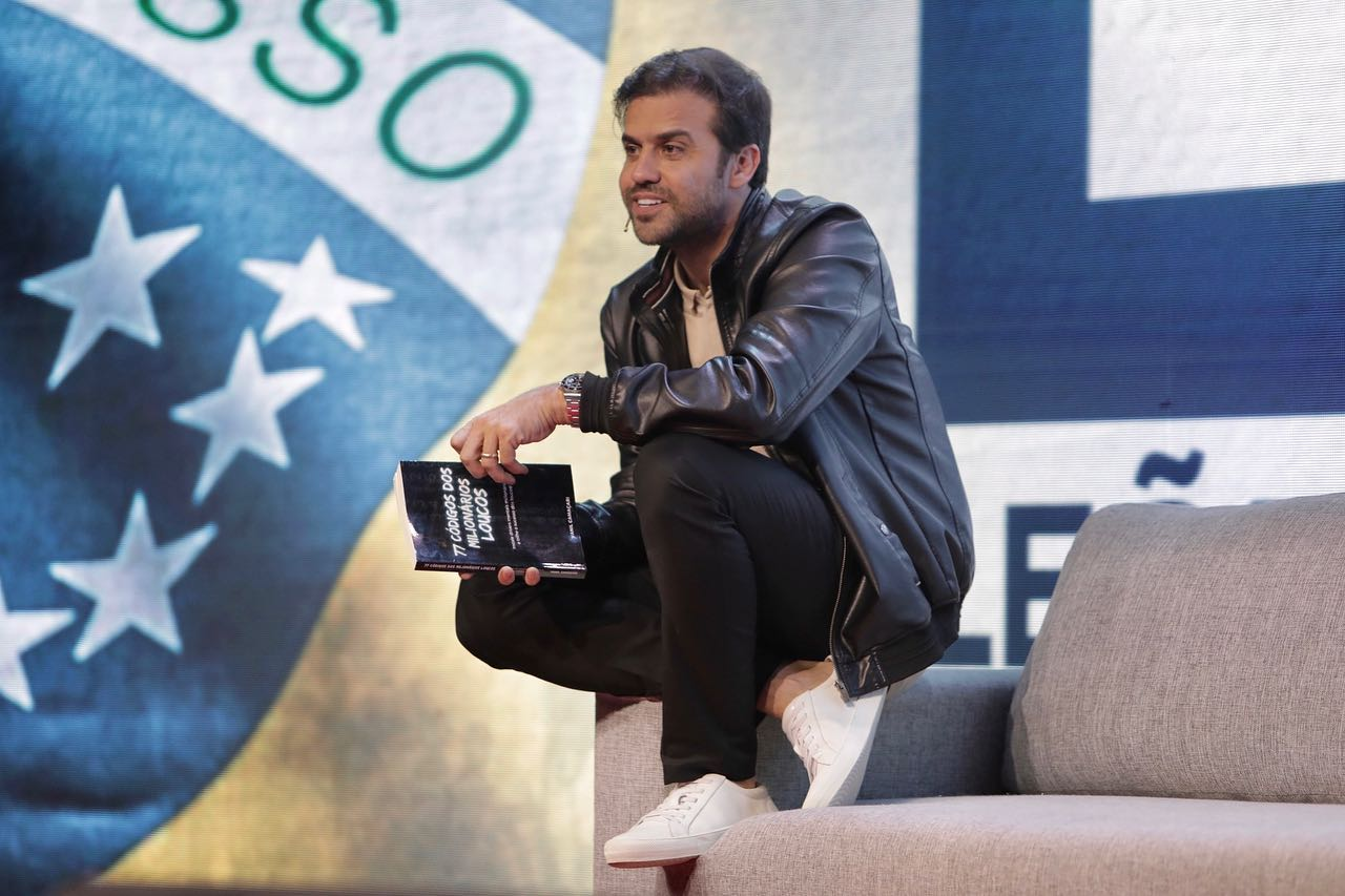 Pablo Marçal (PRTB) sorrindo, agachado, com livro na mão, sem olhar para a câmera, apoiado em poltrona