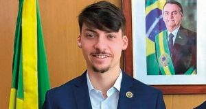 Jair Renan Bolsonaro (PL), candidato a vereador em Balneário Camboriú, em Santa Catarina, sorrindo para a câmera perto de foto do pai e bandeira