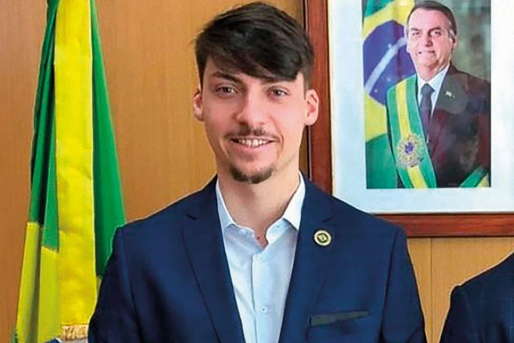 Jair Renan Bolsonaro (PL), candidato a vereador em Balneário Camboriú, em Santa Catarina, sorrindo para a câmera perto de foto do pai e bandeira
