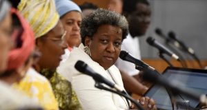 Ministra Macaé Evaristo séria, acompanhada em bancada, olhando para o lado, perto de microfones