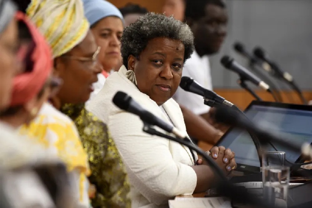 Ministra Macaé Evaristo séria, acompanhada em bancada, olhando para o lado, perto de microfones