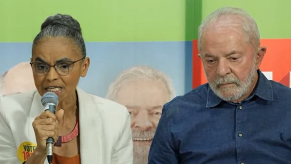 Marina Silva e Lula sentados lado a lado, ela falando em microfone e ele cabisbaixo