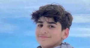 Ali Kamal Abdallah sorrindo de maneira tímida para a câmera com céu azul ao fundo e casaco cinza e vermelho