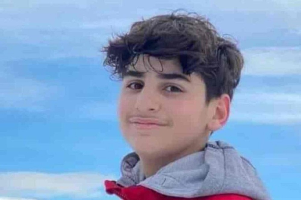 Ali Kamal Abdallah sorrindo de maneira tímida para a câmera com céu azul ao fundo e casaco cinza e vermelho