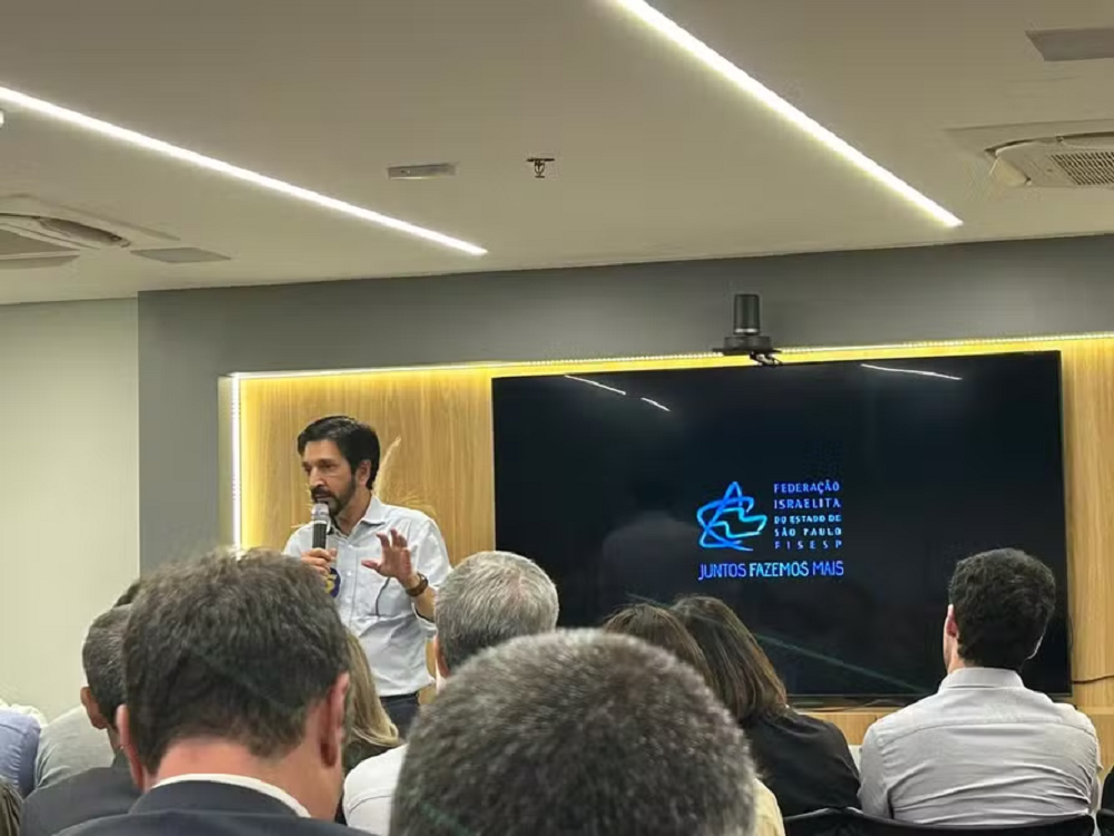Ricardo Nunes (MDB) na Federação Israelista de São Paulo, em pé, falando em microfone e gesticulando, de roupa social clara, sem olhar para a câmera