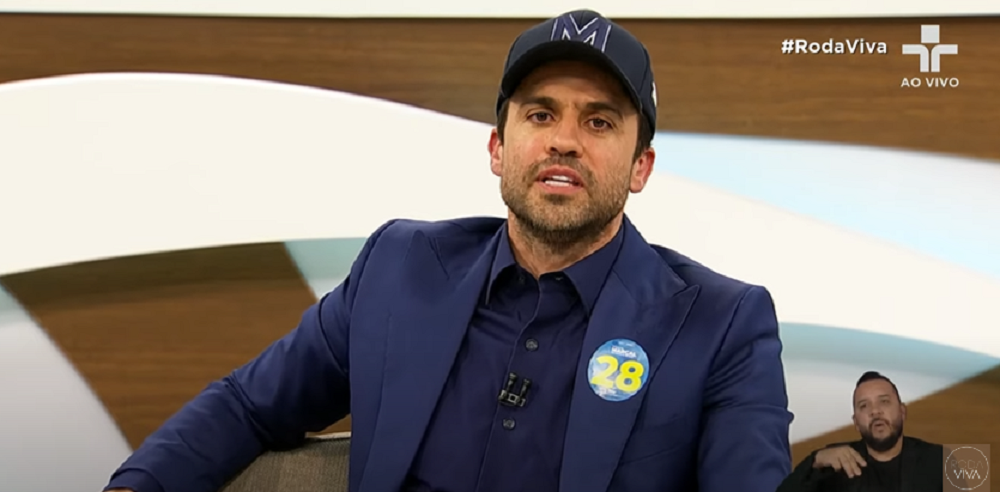 Pablo Marçal no Roda Viva, de roupa azul escura e boné, falando para a câmera com expressão séria